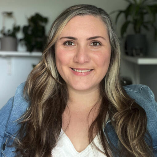 Portrait of Bec Fazzalari, a white woman with long wavy brown hair with blonde highlights wearing a blue denim jacket over white top, smiling with houseplants in the background.